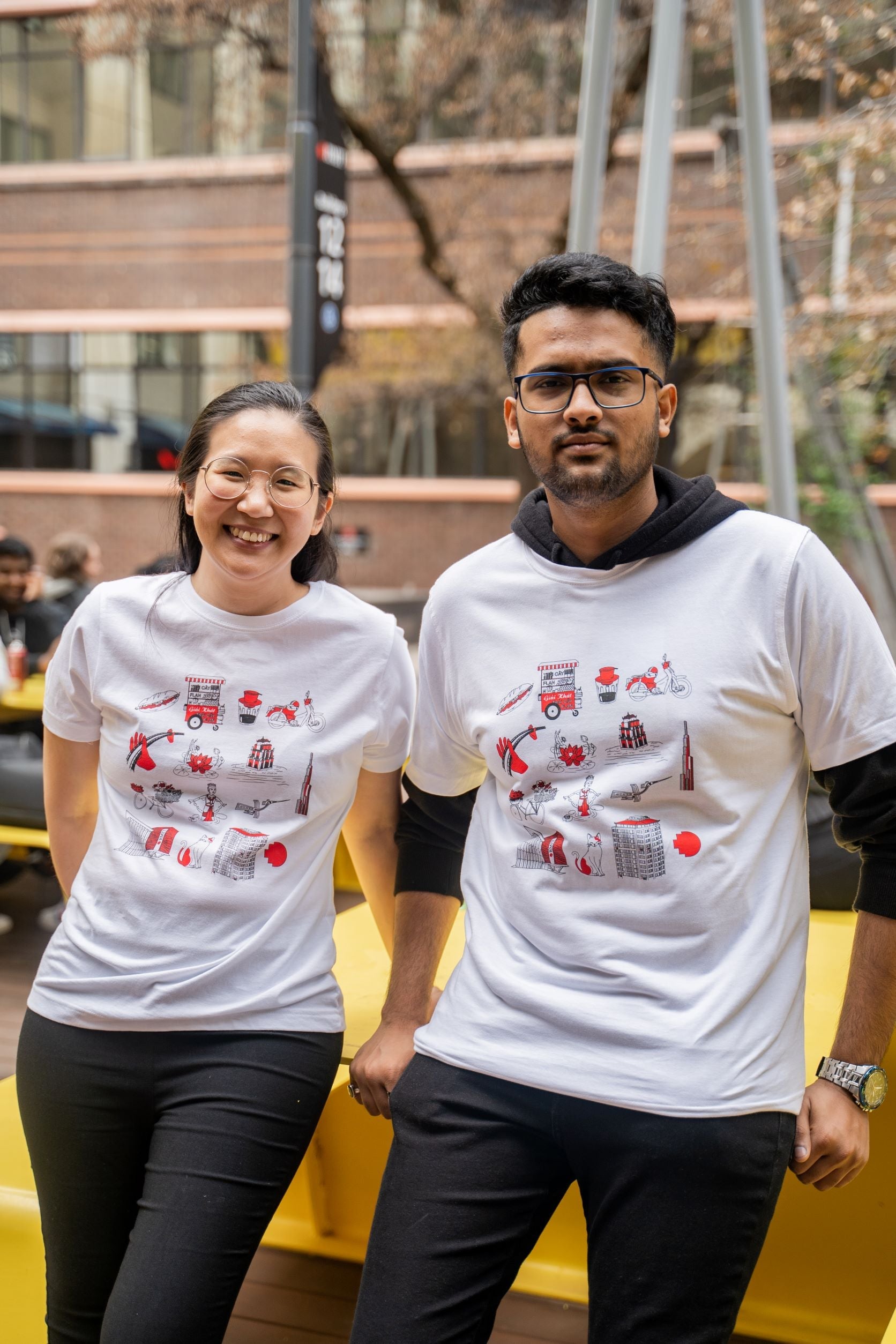 Two models wearing the Vietnamese Icon Tee from RMIT Store sitting against a yellow bench. The white T-shirt features red and black graphics inspired by Vietnamese culture, including traditional food carts and iconic buildings.